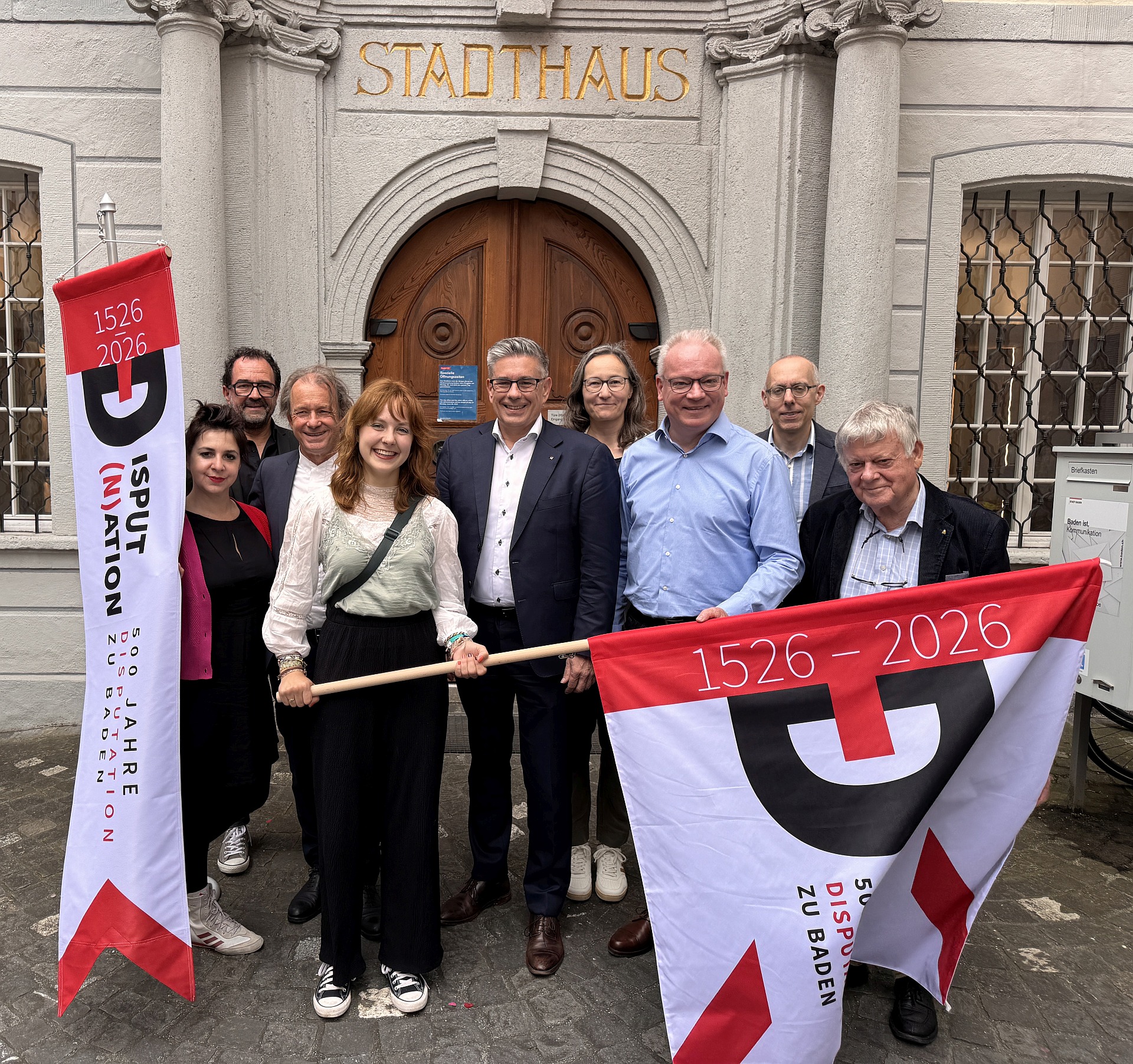 Mitglieder der Organisation u.a. mit Res Peter, ref. Pfarrer, Vorsitzender der Projektleitung (3.vl), Claudio Tomassini, kath. Seelsorger und Pfarreileiter (3. vr), Markus Schneider. Stadtammann von Baden (Mitte) und der Jubla-Vertreterin, die die Fahne ins Jublasurium getragen hat